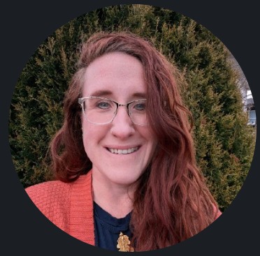 A red-haired woman with gold glasses and an orange shrug smiles in front of an evergreen tree